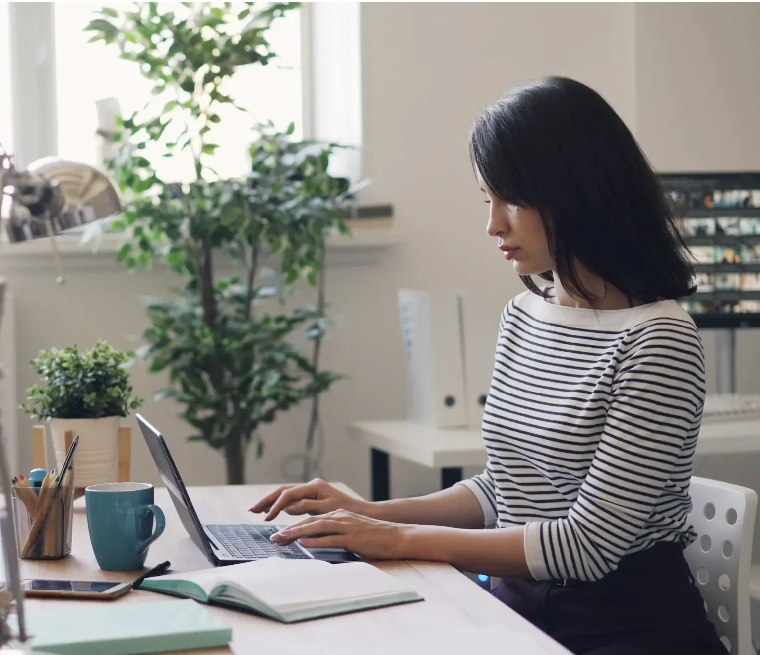 Designer working at a sunlit desk with a laptop, notebook, and coffee